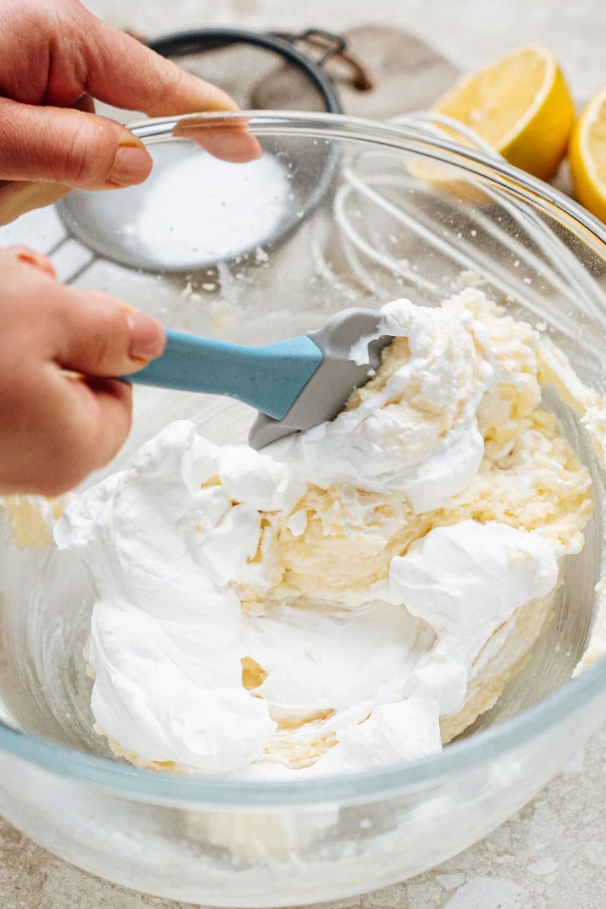 Hands mixing whipped cream with a spatula in a glass bowl, with lemon halves and a sifter in the background.