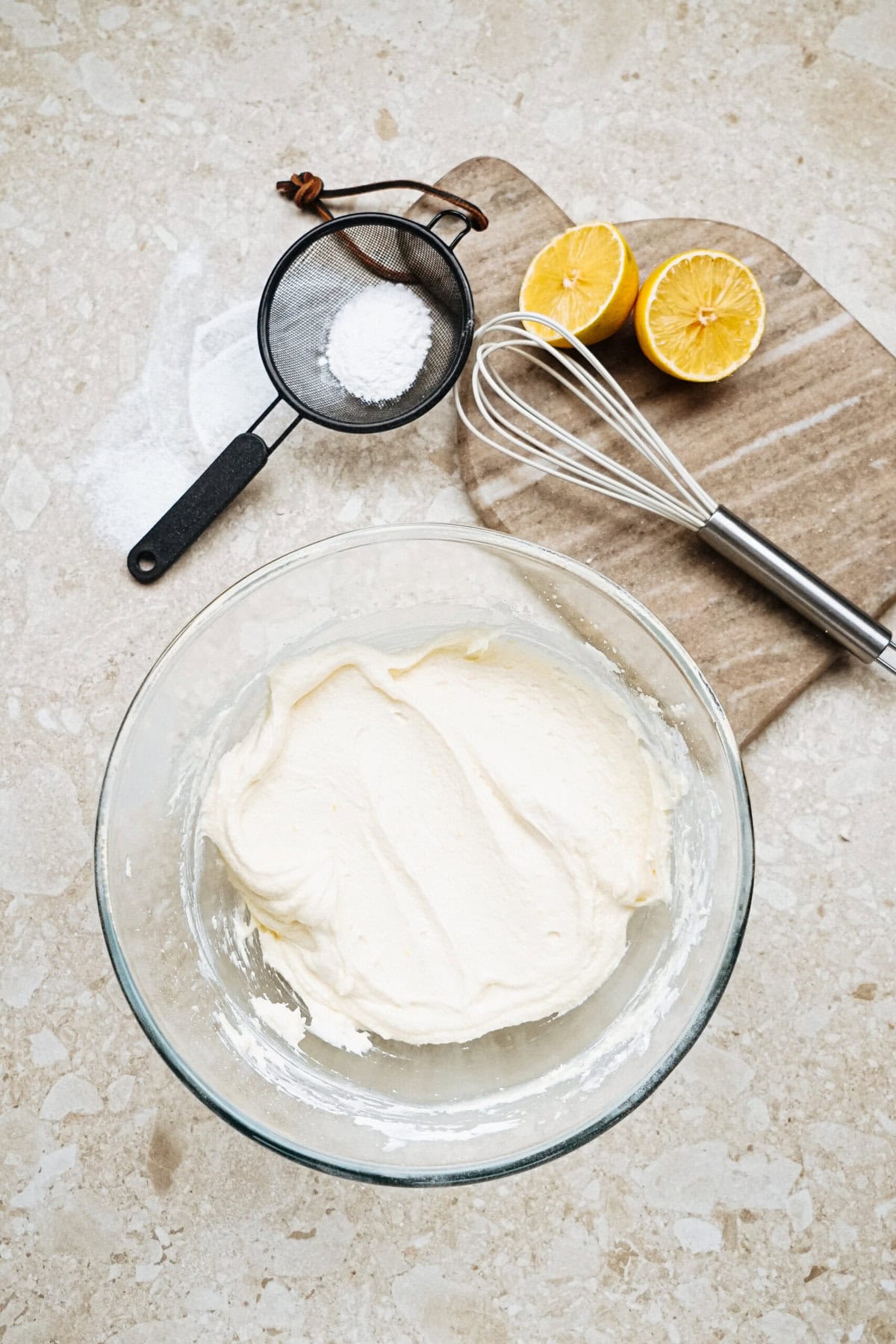 A bowl of creamy batter on a countertop, with a whisk, halved lemon, and a sifter containing flour nearby.