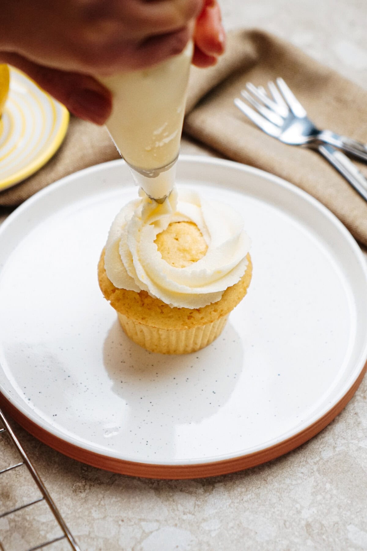 Person decorating a cupcake with white frosting on a white plate.