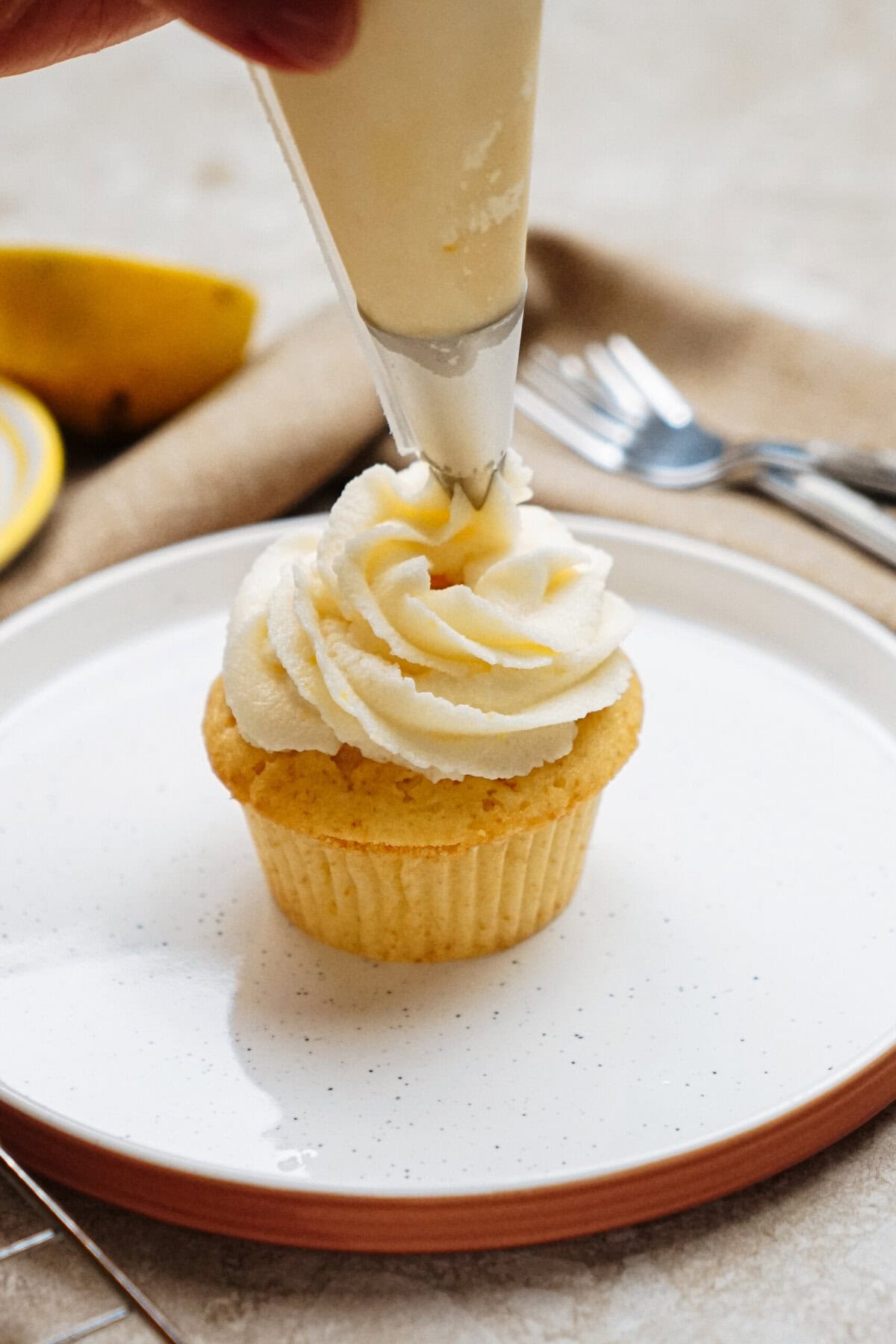 A hand pipes frosting onto a cupcake on a white plate, with a fork and a lemon slice in the background.