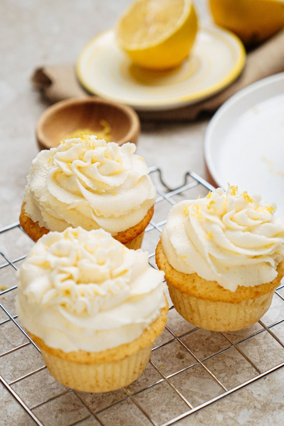 Three lemon cupcakes with creamy frosting sit on a cooling rack. A halved lemon is in the background on a plate.