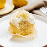 A partially eaten lemon cupcake with creamy frosting on a plate, surrounded by lemon halves and a fork.