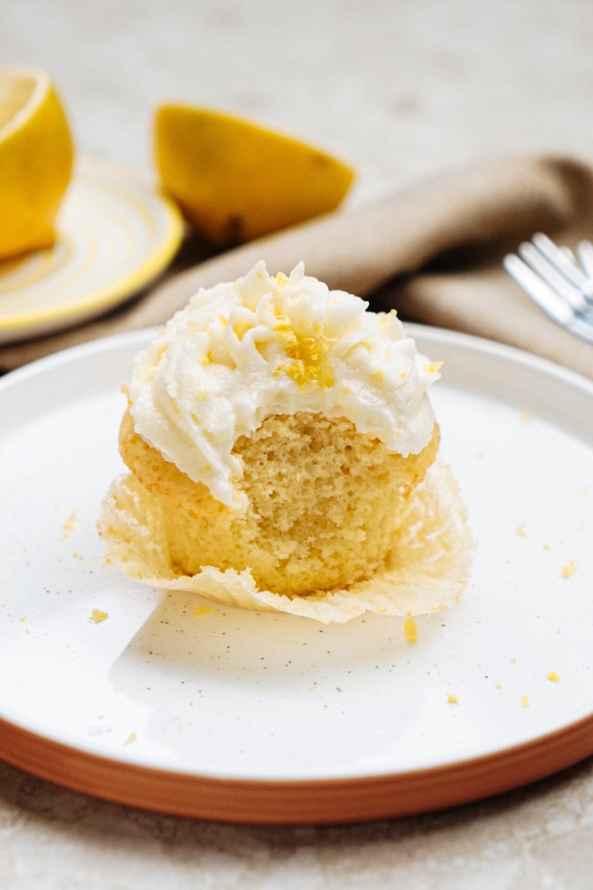 A partially eaten lemon cupcake with creamy frosting on a plate, surrounded by lemon halves and a fork.