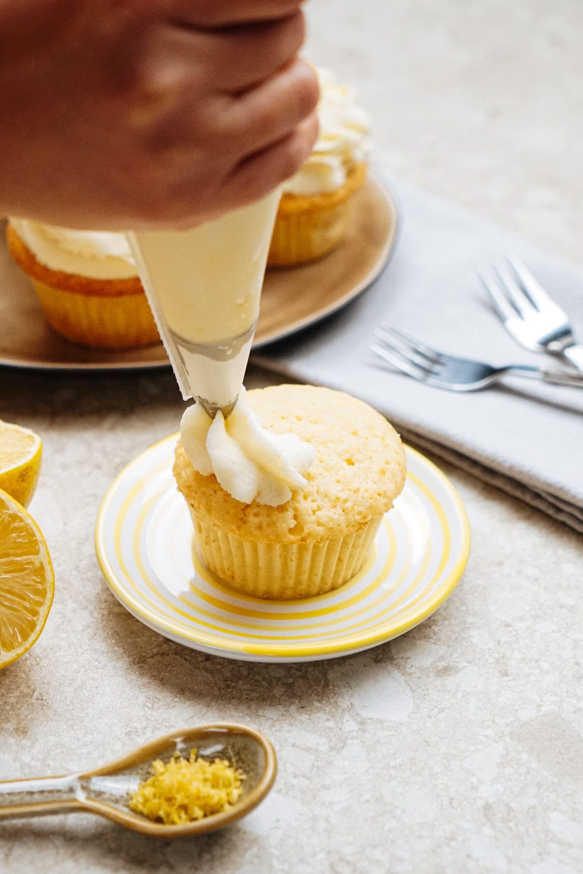 A hand is piping cream onto a cupcake on a striped plate. Lemons, lemon zest, and additional cupcakes are visible in the background.