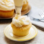 A person piping frosting onto a cupcake on a yellow-striped plate, with more cupcakes and utensils in the background.