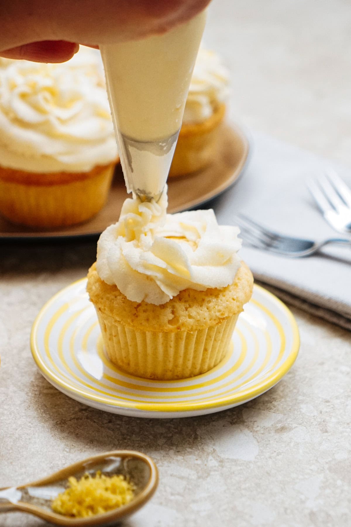 A person piping frosting onto a cupcake on a yellow-striped plate, with more cupcakes and utensils in the background.