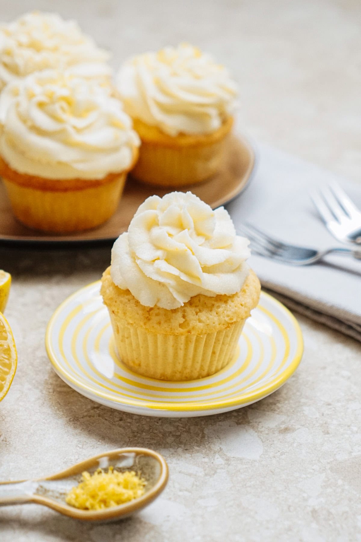 A vanilla cupcake with white frosting on a striped plate. More cupcakes are on a tray in the background, with lemon zest and utensils nearby.