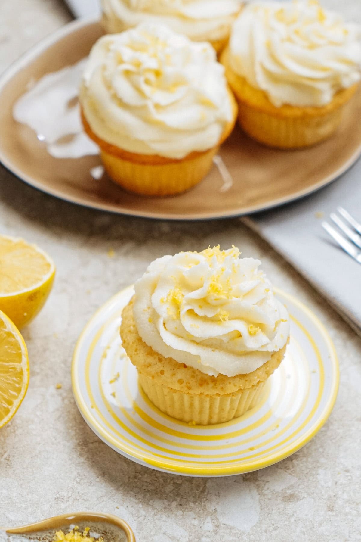 A lemon cupcake with frosting and lemon zest sits on a striped plate. In the background, more cupcakes and lemon halves are visible.