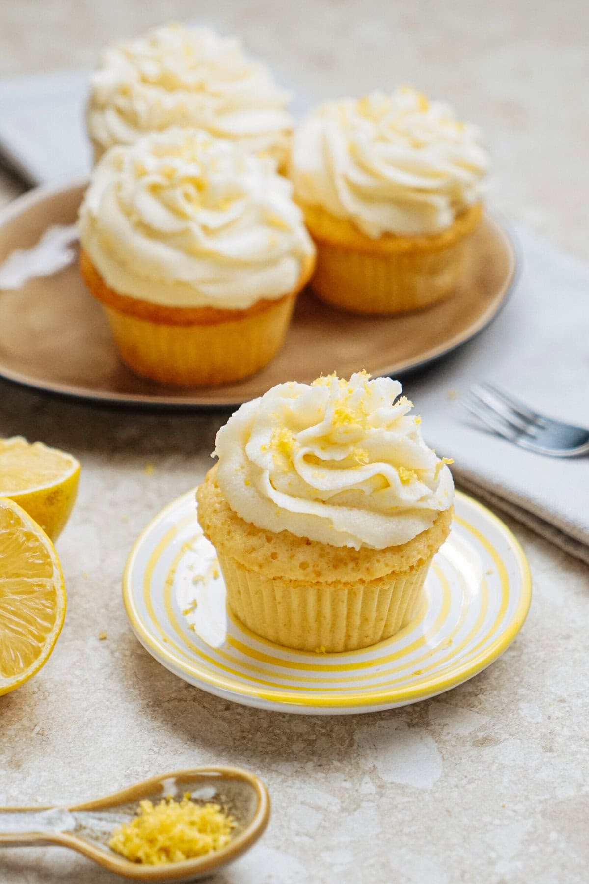 Lemon cupcakes with creamy frosting on plates; one is in the foreground on a striped plate with lemon zest, a lemon half, and a spoonful of zest nearby.