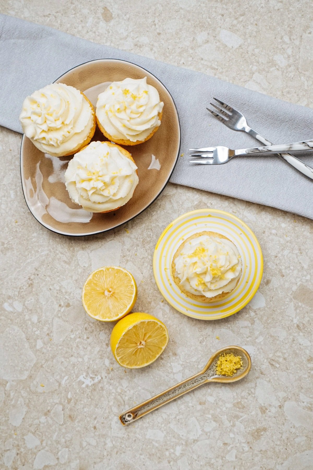 Cupcakes with white frosting and lemon zest on plates, accompanied by halved lemons, a spoon of zest, and forks on a gray napkin.