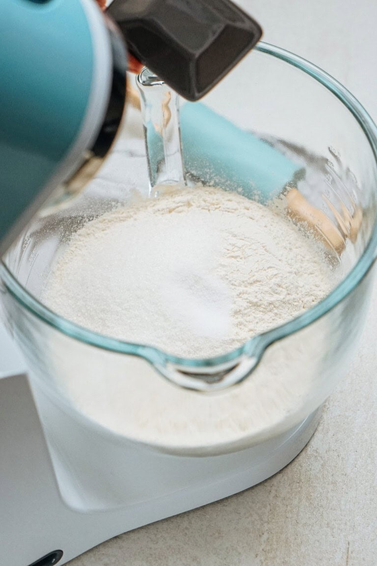 A stand mixer with a glass bowl contains flour and sugar being mixed.