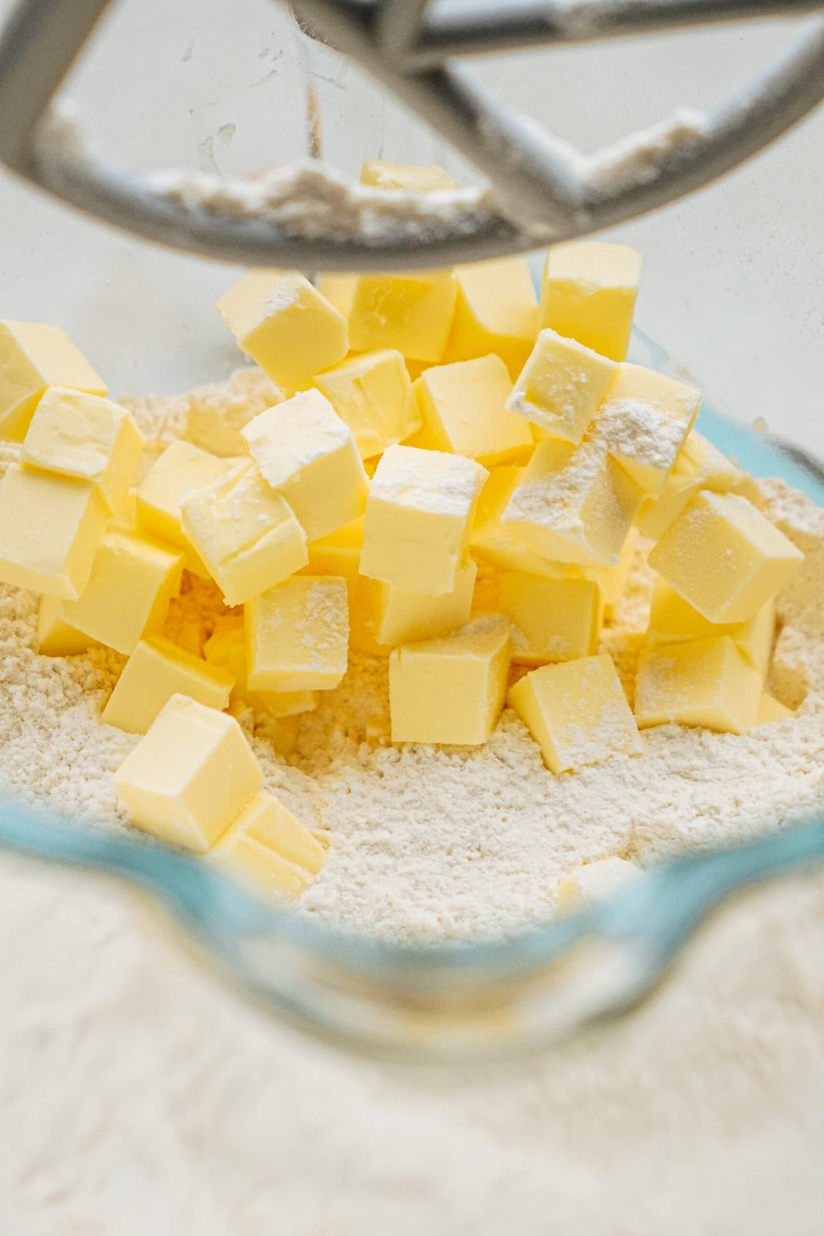 Close-up of cubed butter mixed with flour in a glass bowl, with a metal mixer attachment visible above.