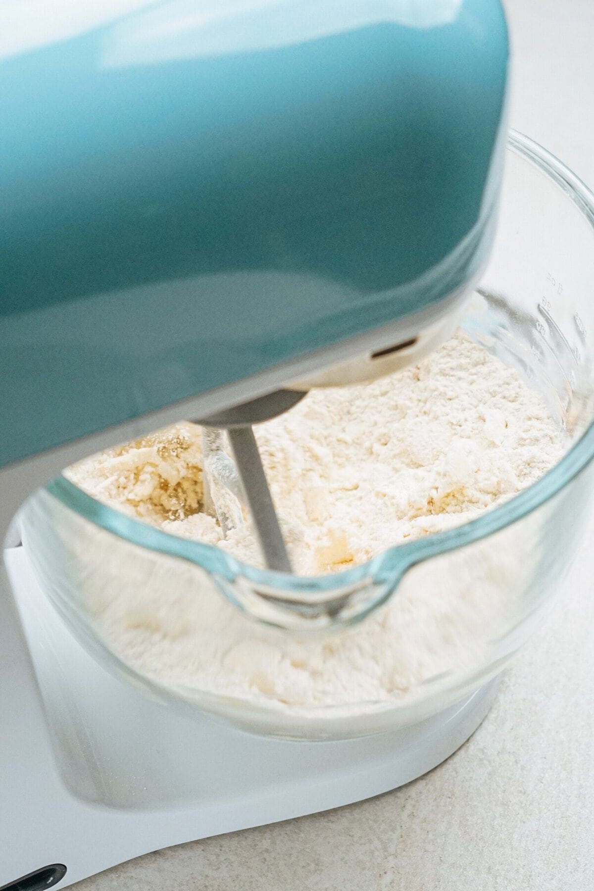 A stand mixer with a blue top is mixing flour in a clear glass bowl on a kitchen countertop.