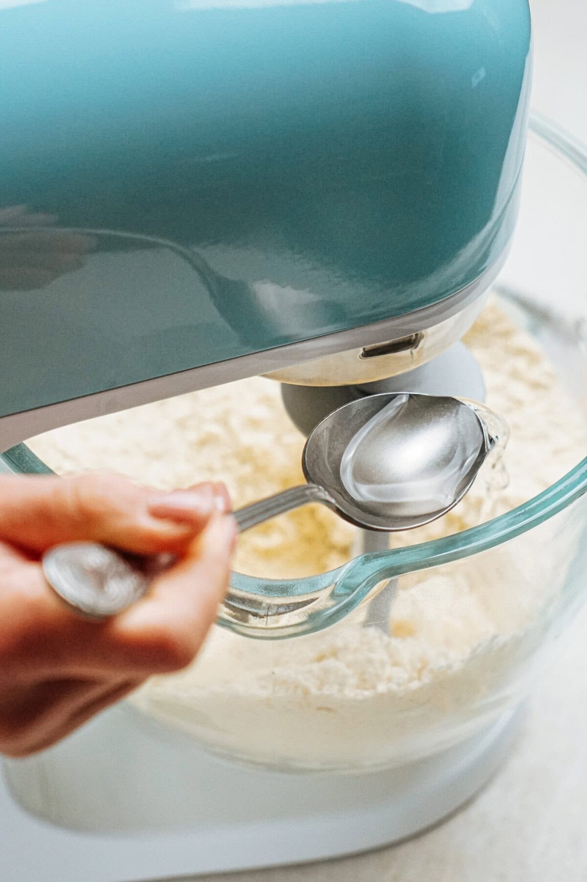 A person adds liquid from a spoon into a blue stand mixer with a glass bowl containing dry ingredients.