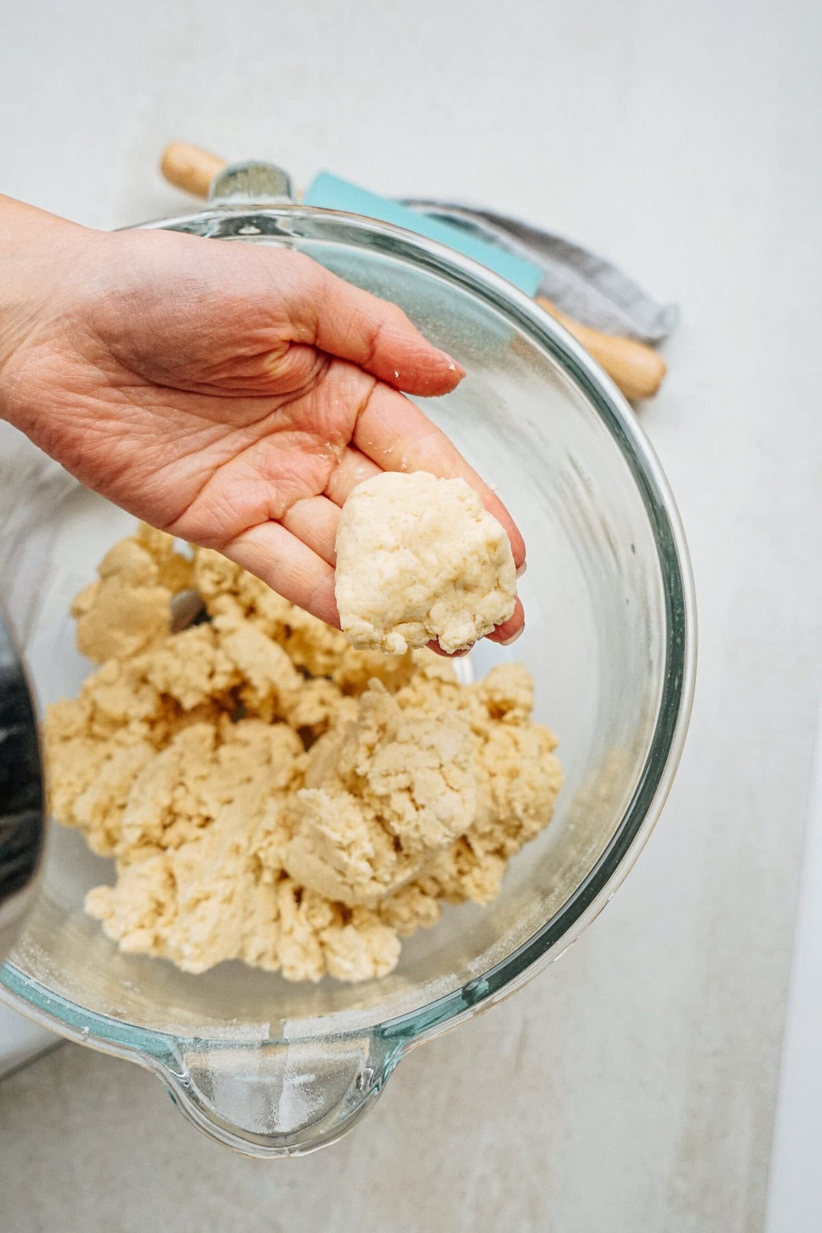 A hand holds a piece of dough over a glass bowl filled with more dough. A rolling pin is visible in the background.