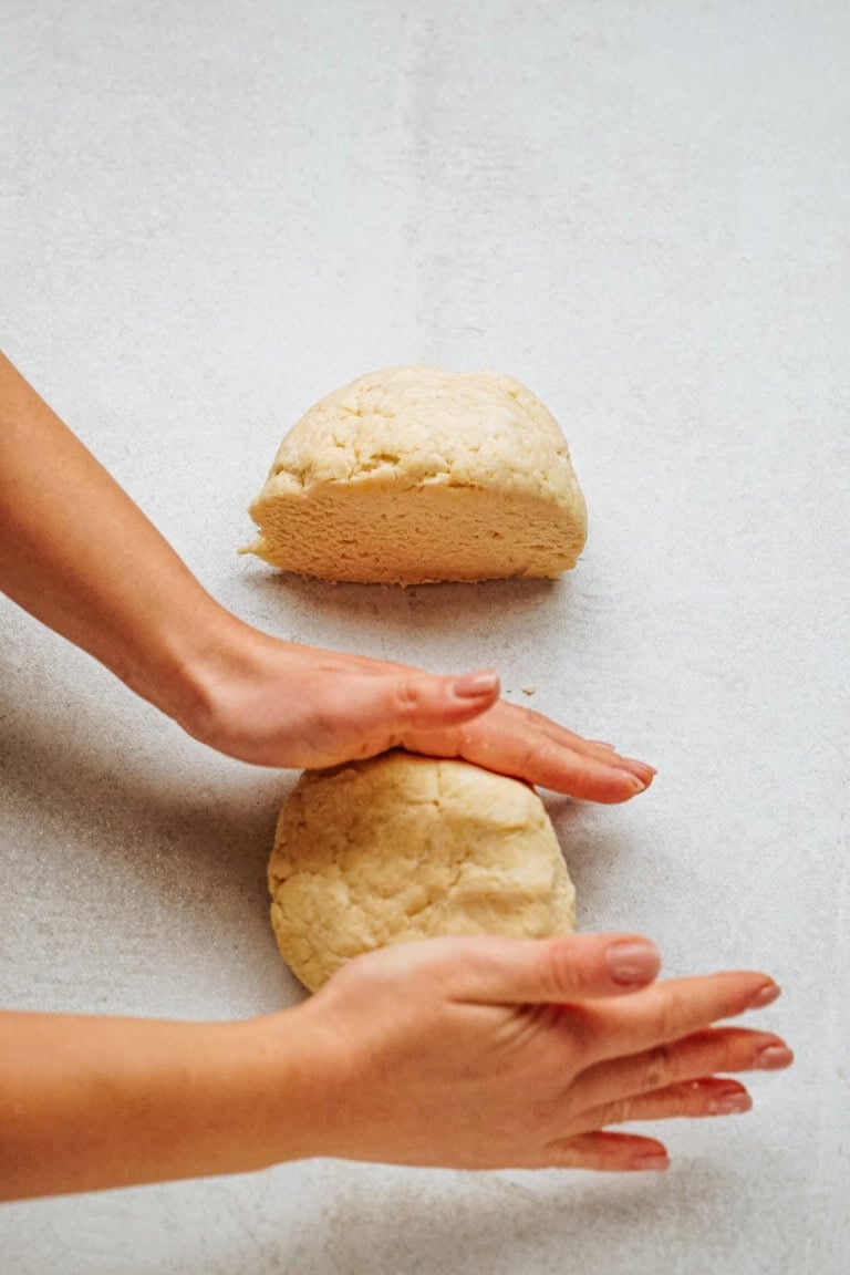 Hands shaping two pieces of dough on a light surface.