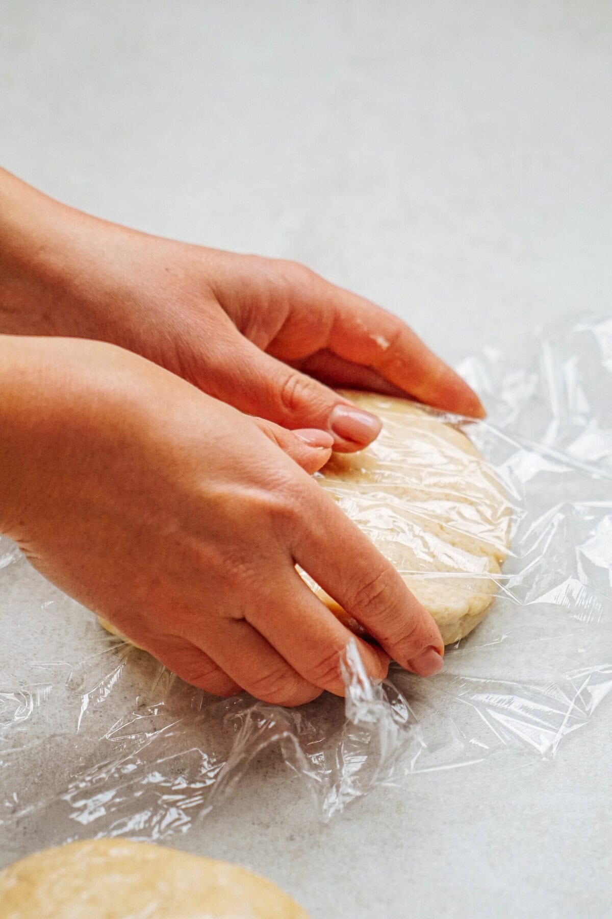 Hands wrapping dough in plastic wrap on a light-colored surface.