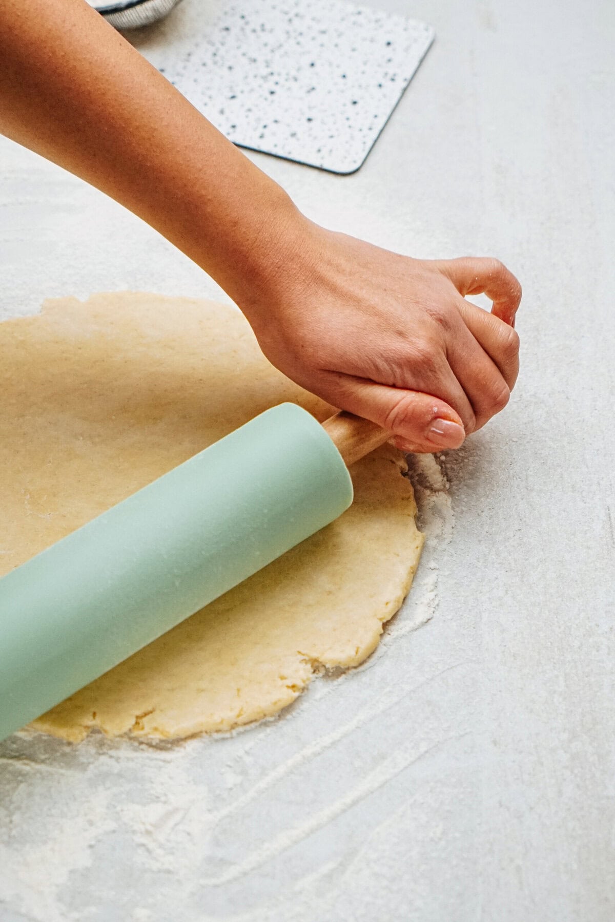 A hand using a green rolling pin to flatten dough on a floured surface.