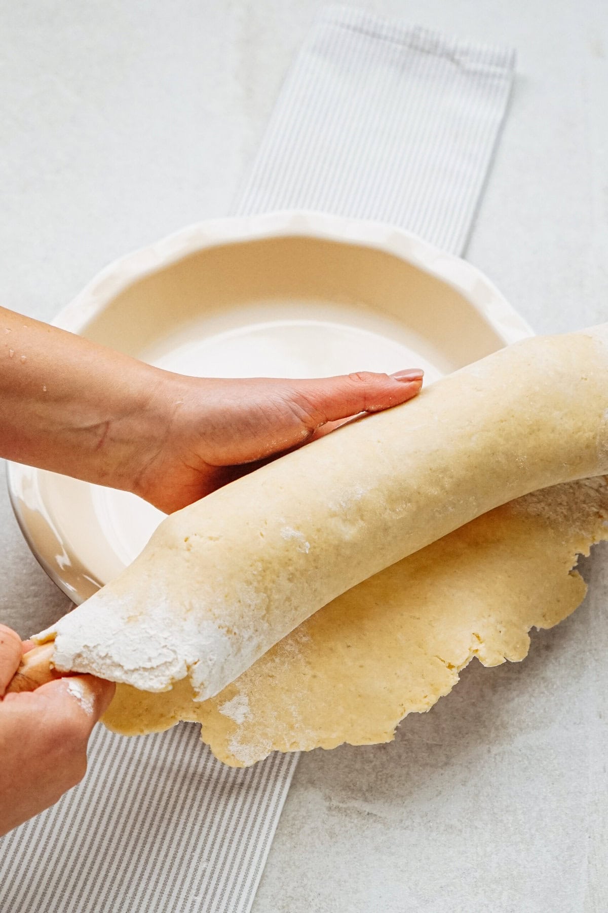 Person placing rolled-out dough into a pie dish on a striped cloth surface.