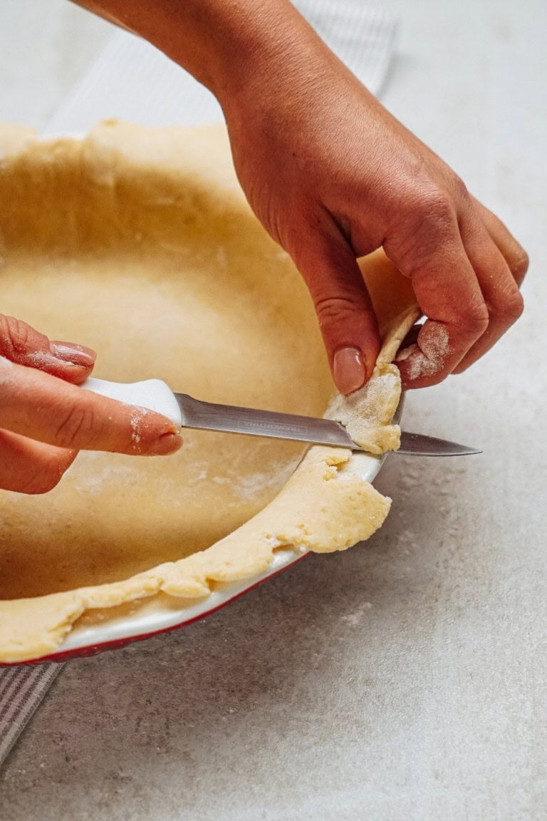 Hands trimming excess dough from a pie crust in a dish with a small knife on a light-colored surface.