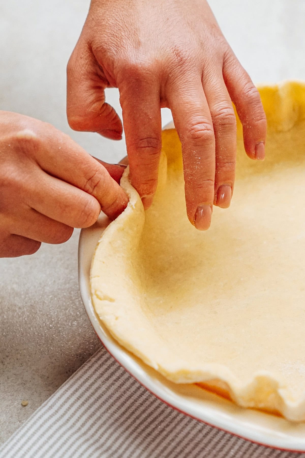 Hands are crimping the edge of an unbaked pie crust in a round dish on a striped cloth.