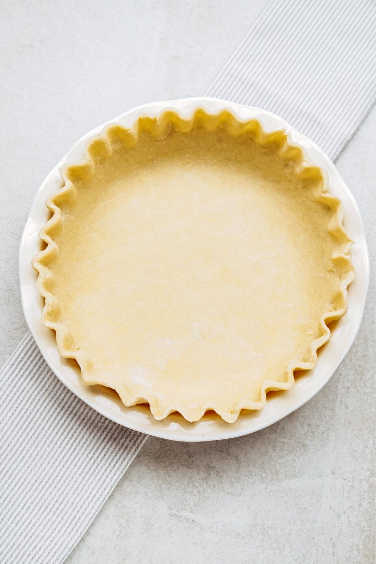 A pie crust with crimped edges in a white pie dish on a gray-striped surface.