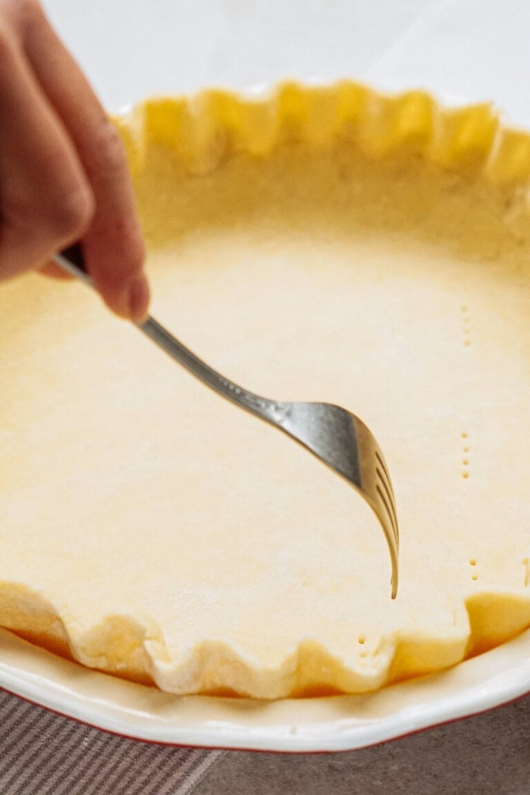 A hand uses a fork to prick holes in an uncooked pie crust in a pan.