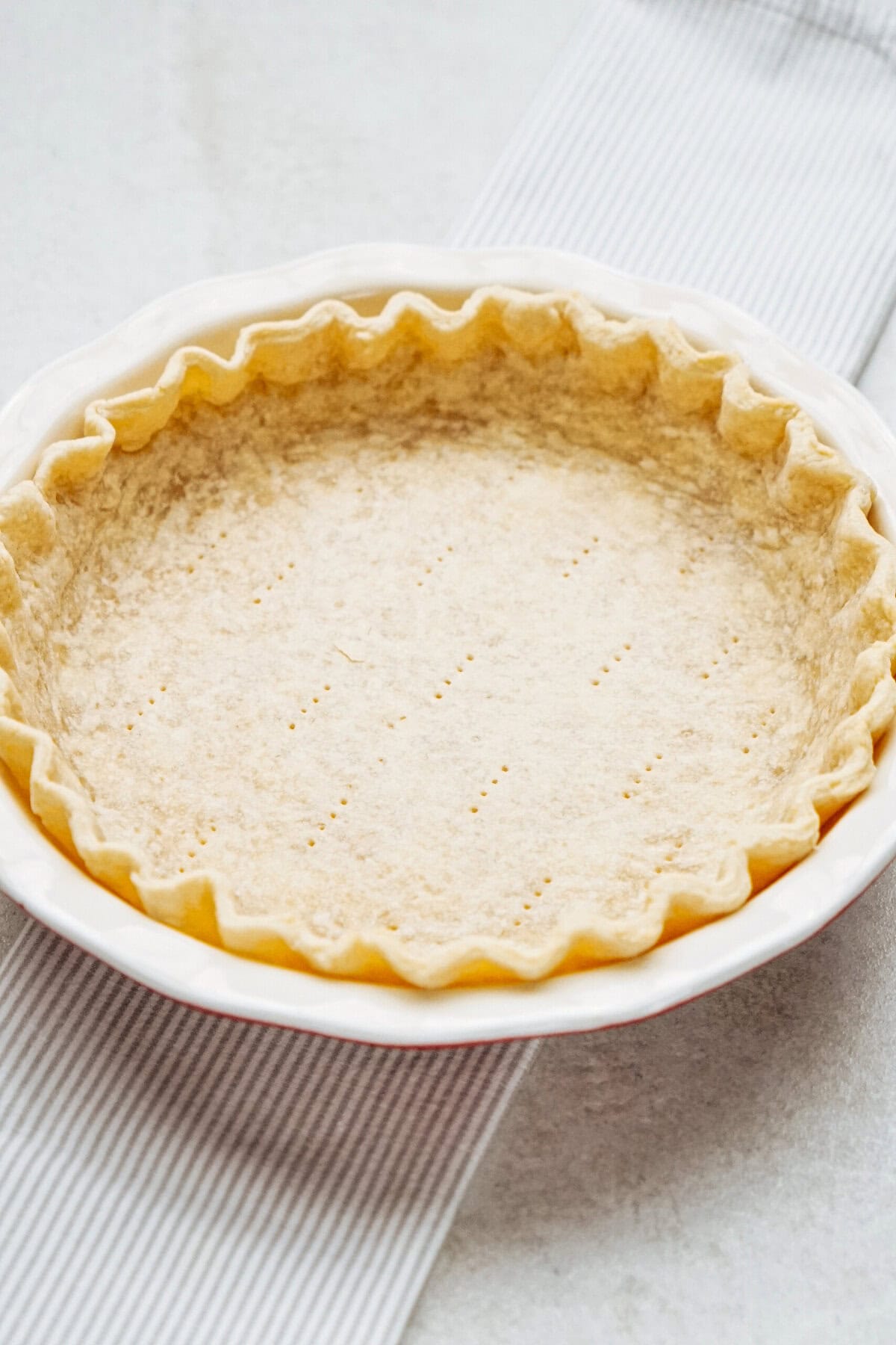 Unbaked pie crust with fluted edges in a white pie dish, resting on a striped fabric.