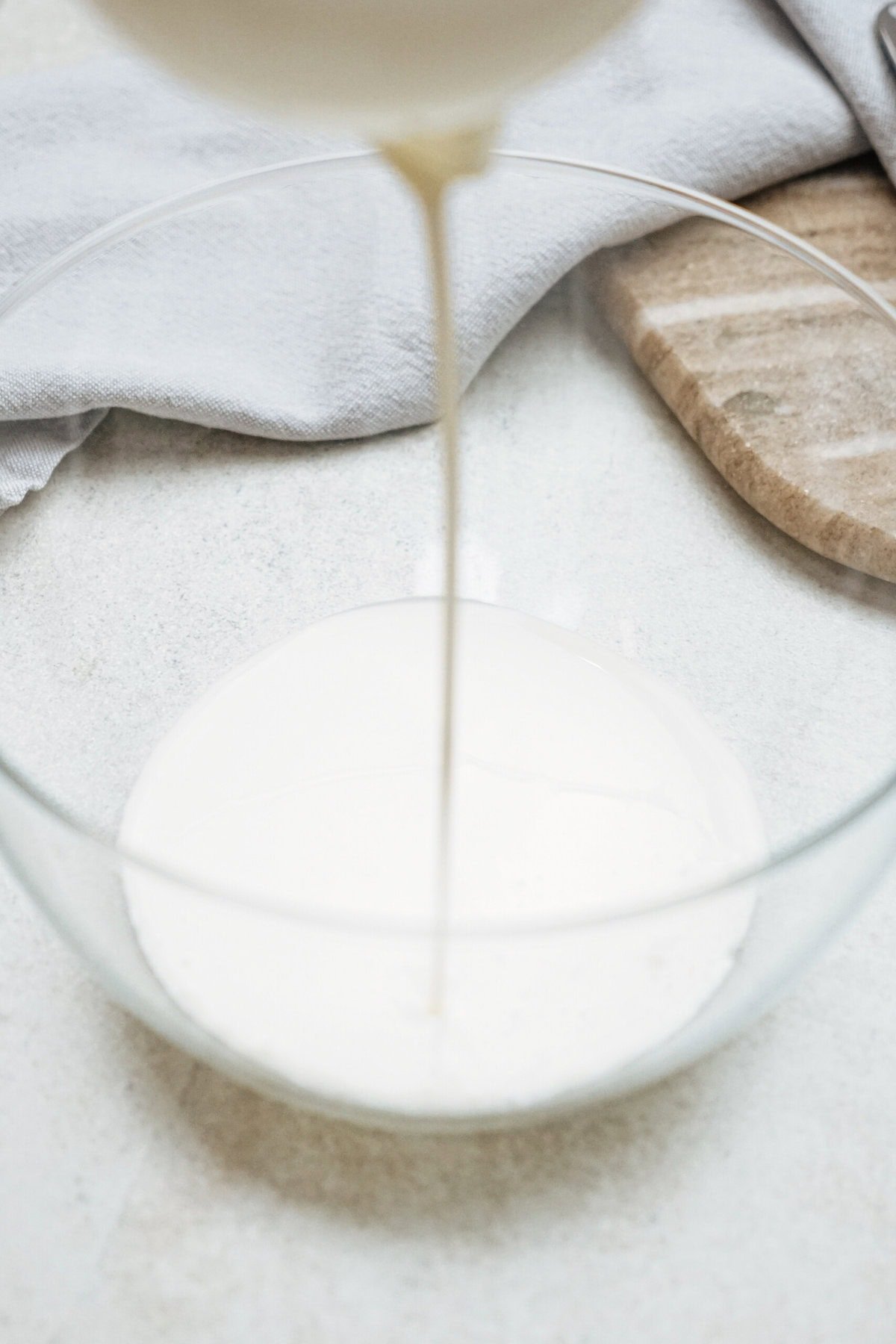 Heavy cream being poured into a clear glass bowl on a light countertop, with a grey cloth and wooden board in the background.