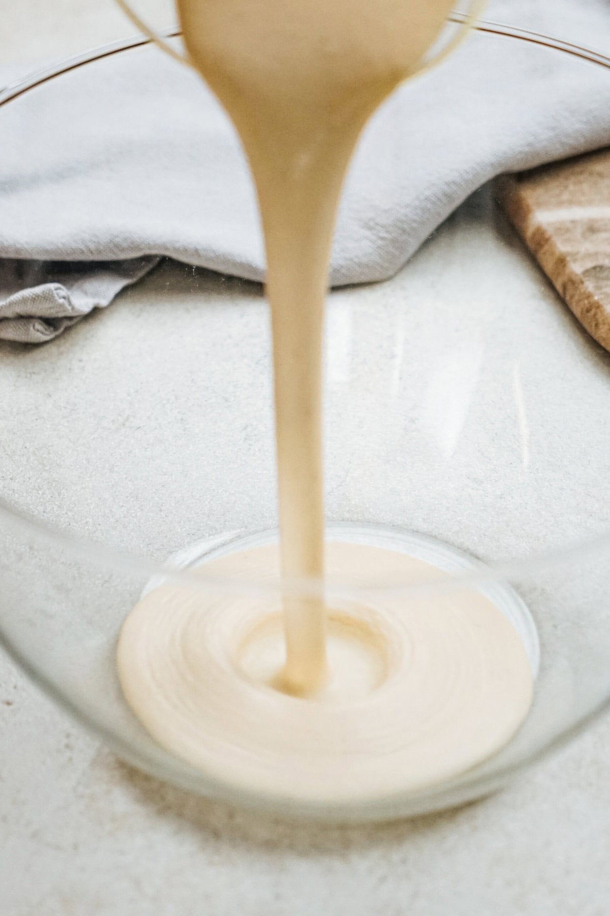 Batter being poured into a clear glass bowl on a white surface, with a folded cloth and wooden board nearby.