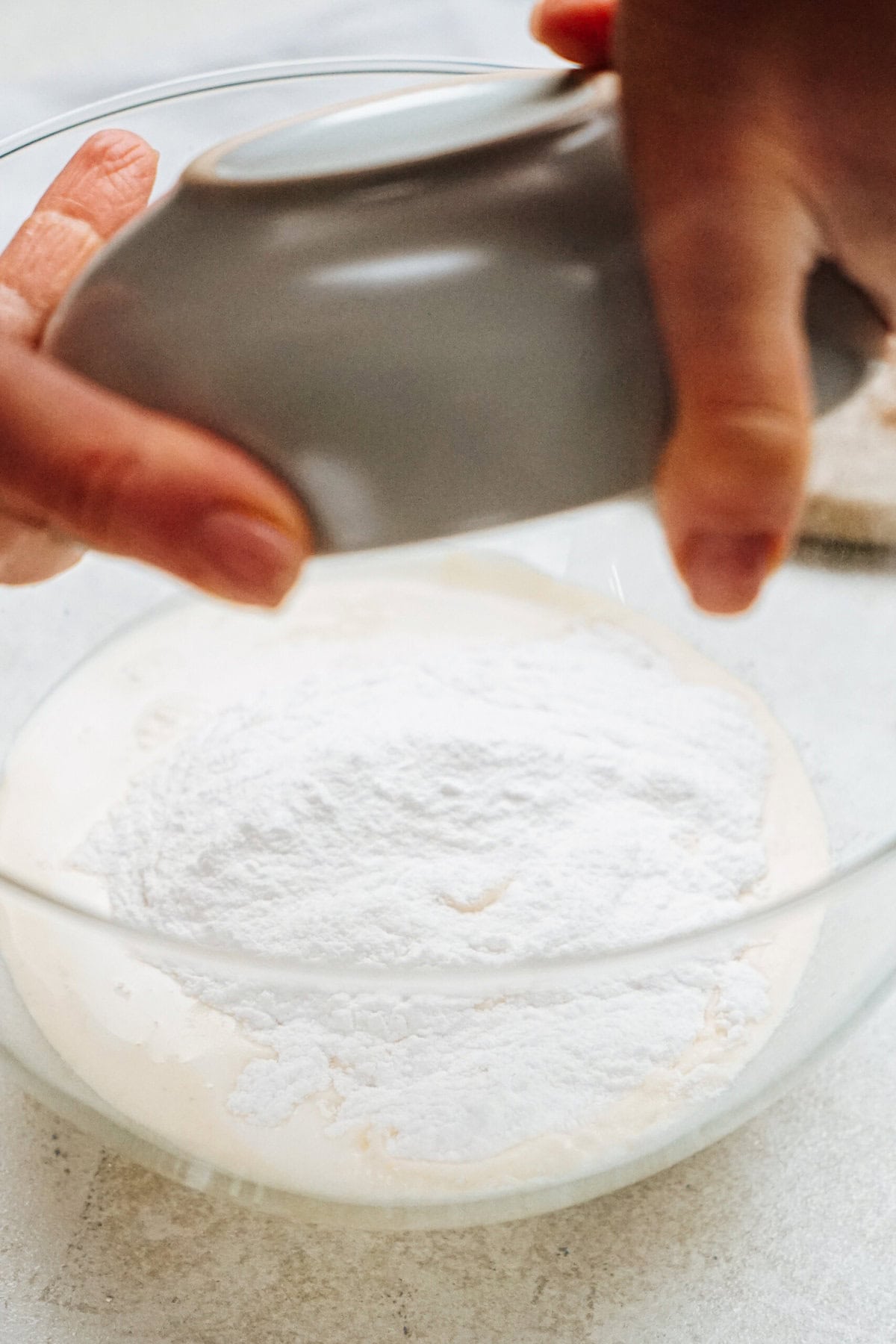 Hands pouring flour from a gray bowl into a glass mixing bowl.