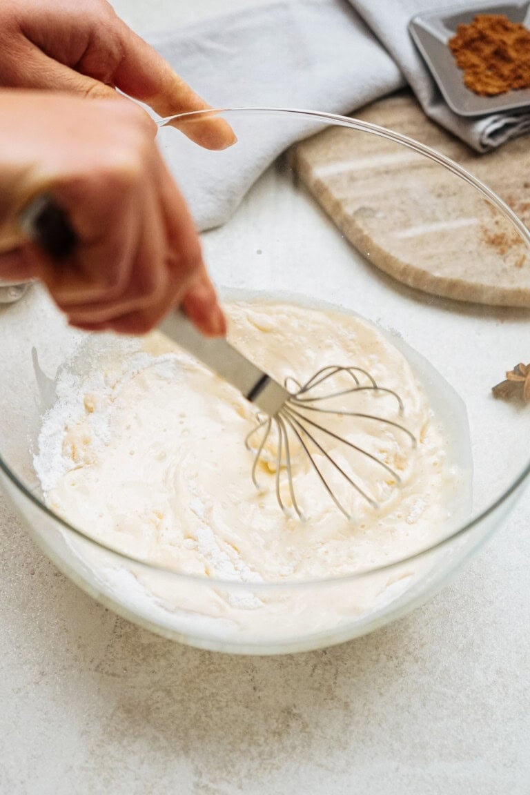 A person whisking a creamy mixture in a glass bowl on a white countertop, with a grey napkin and spices in the background.