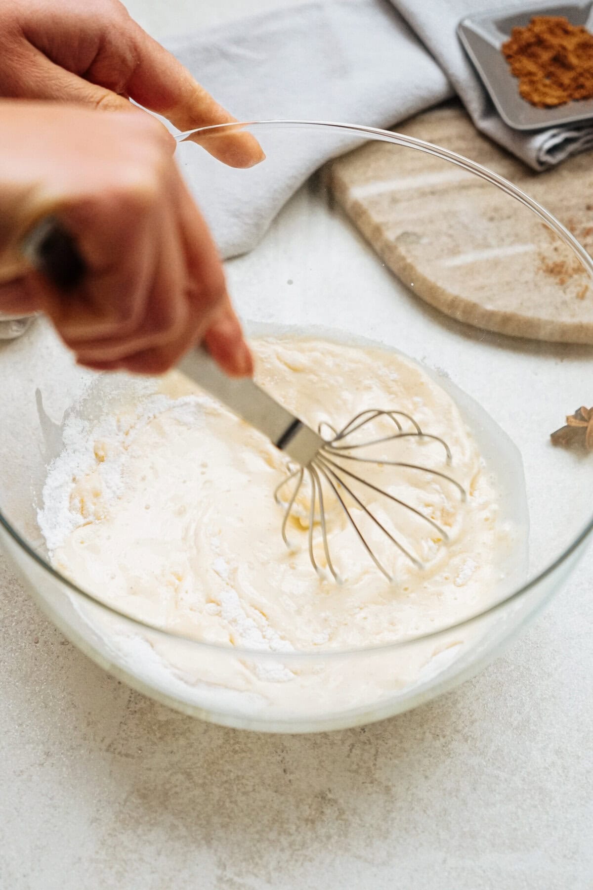 A person whisking a creamy mixture in a glass bowl on a white countertop, with a grey napkin and spices in the background.