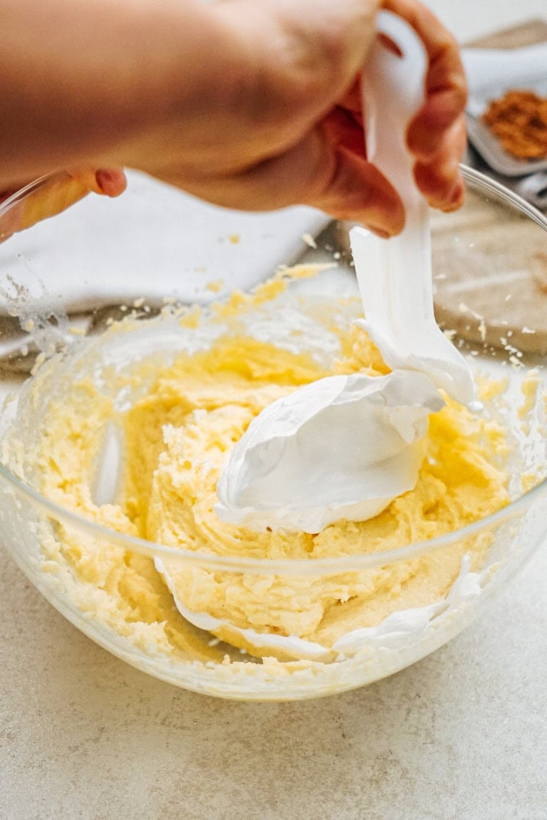 Hands holding a spatula, mixing cream into a bowl of yellow batter on a countertop.