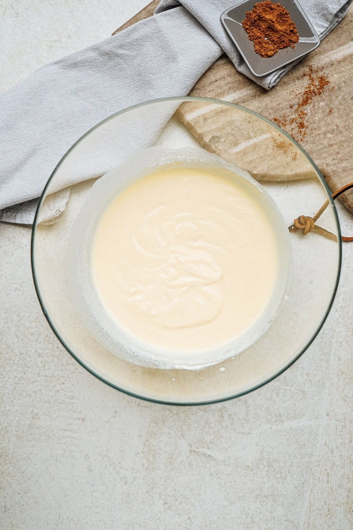 A glass bowl filled with a creamy batter on a light surface, accompanied by a cloth and a small plate with brown spice.