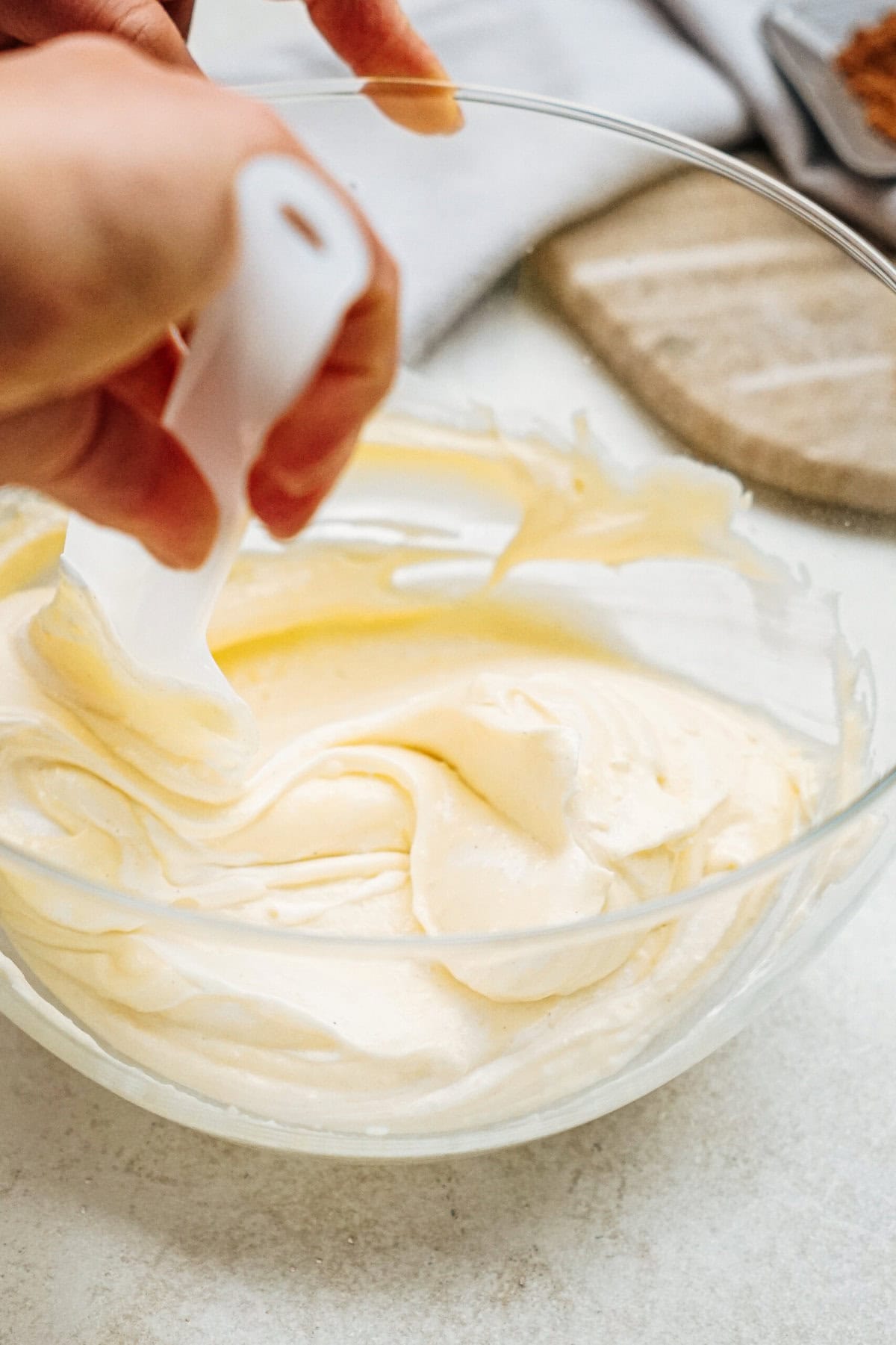 A person uses a spatula to mix creamy batter in a clear glass bowl on a light-colored surface.