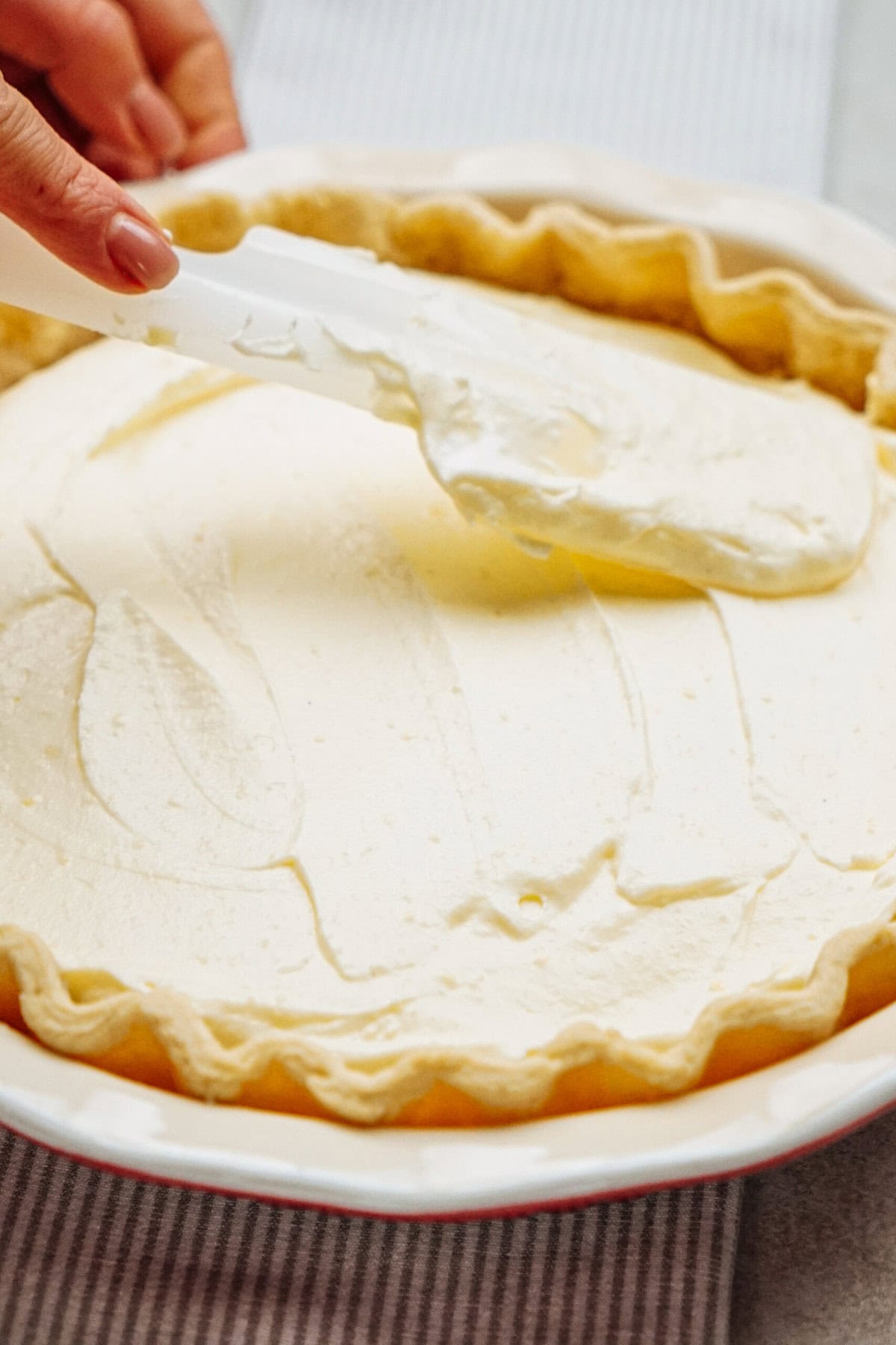 A hand spreads cream filling in a pie crust with a spatula.