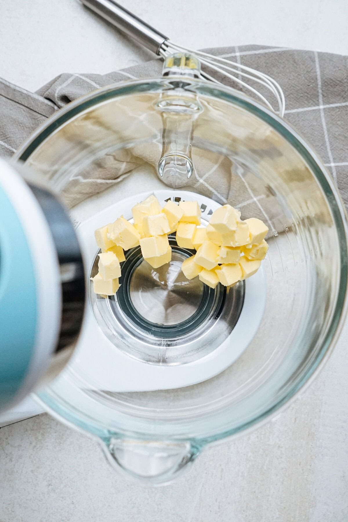 Butter cubes in a glass mixing bowl on a patterned cloth, near a kitchen appliance.