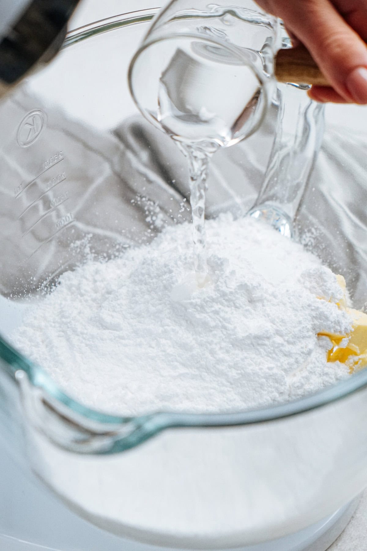 A person pours liquid from a glass pitcher into a bowl with white powder, possibly flour, in a kitchen setting.