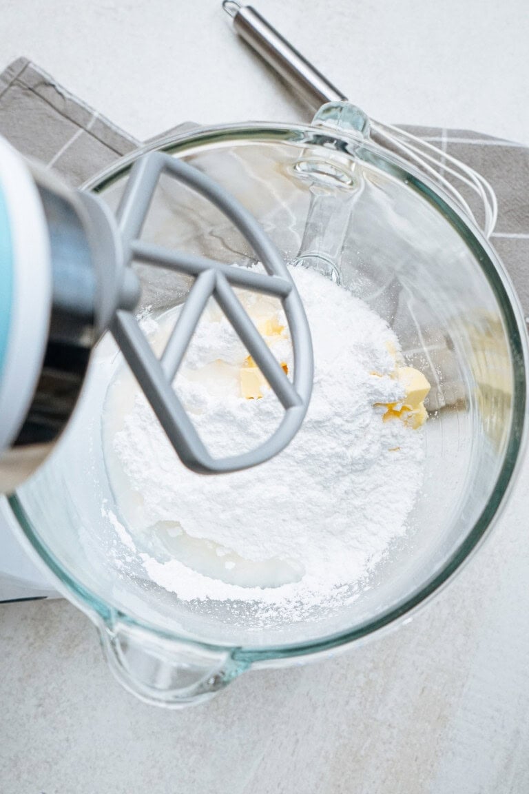 A mixer with a beater attachment is in a glass bowl holding flour, sugar, and butter, ready for mixing on a light-colored countertop.