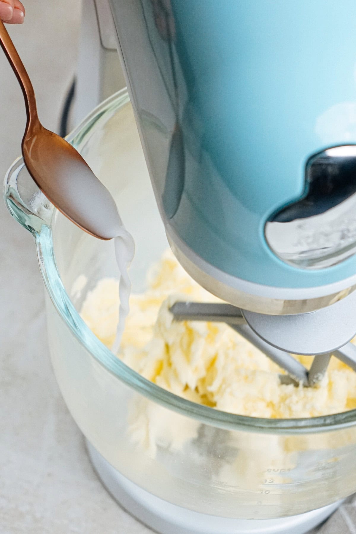 A person adds liquid from a spoon into a stand mixer bowl containing a creamy mixture.