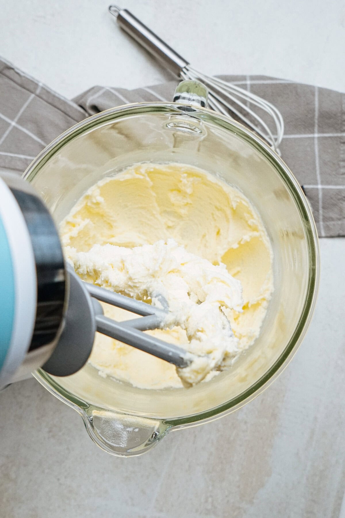 A stand mixer cream paddle mixes butter and sugar in a clear bowl, on a gray checkered cloth with a whisk nearby.