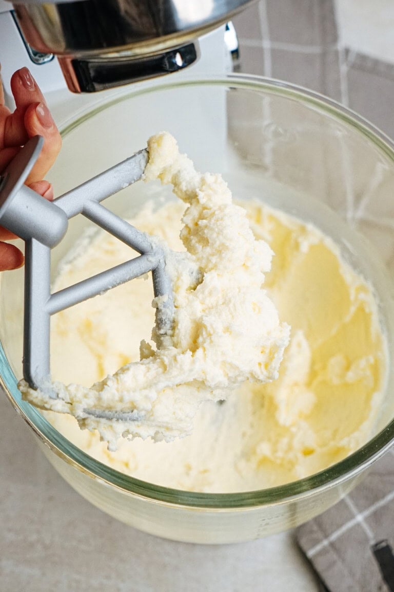 Hand holding a mixer paddle with creamy batter over a glass mixing bowl.