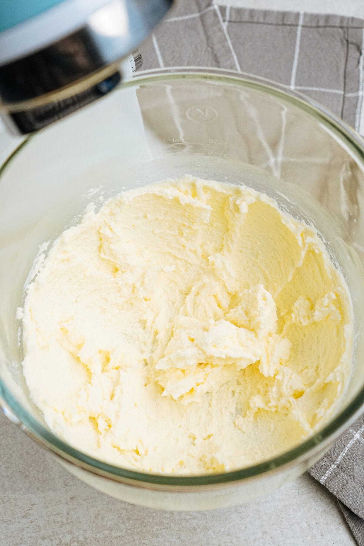 A glass bowl containing creamy, whipped butter and sugar mixture. It is placed on a checkered cloth and part of a mixer is visible above.