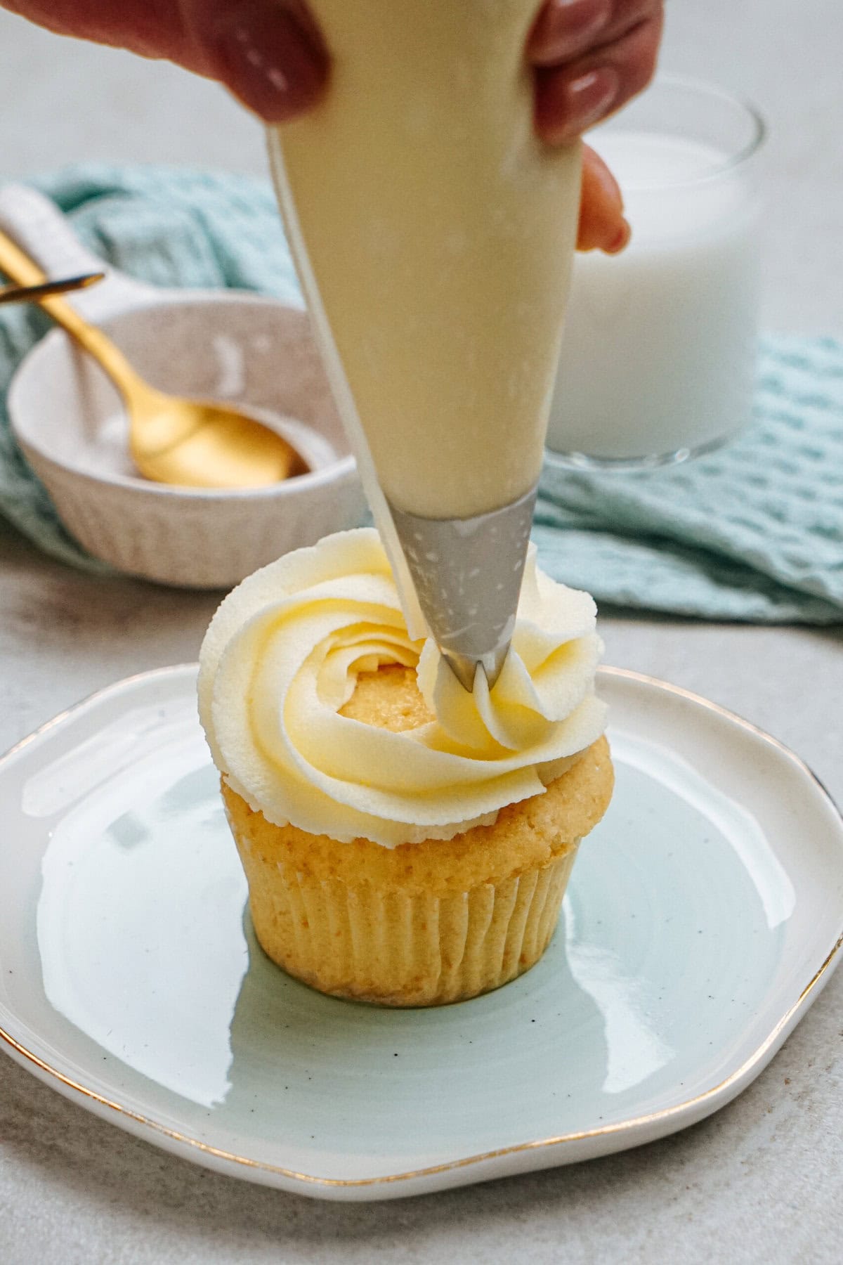 A hand pipes white frosting onto a vanilla cupcake on a plate. A spoon and a bowl are in the background.