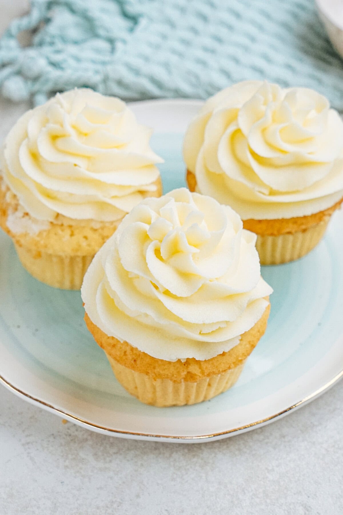 Three vanilla cupcakes with swirls of white frosting on a blue-rimmed plate.