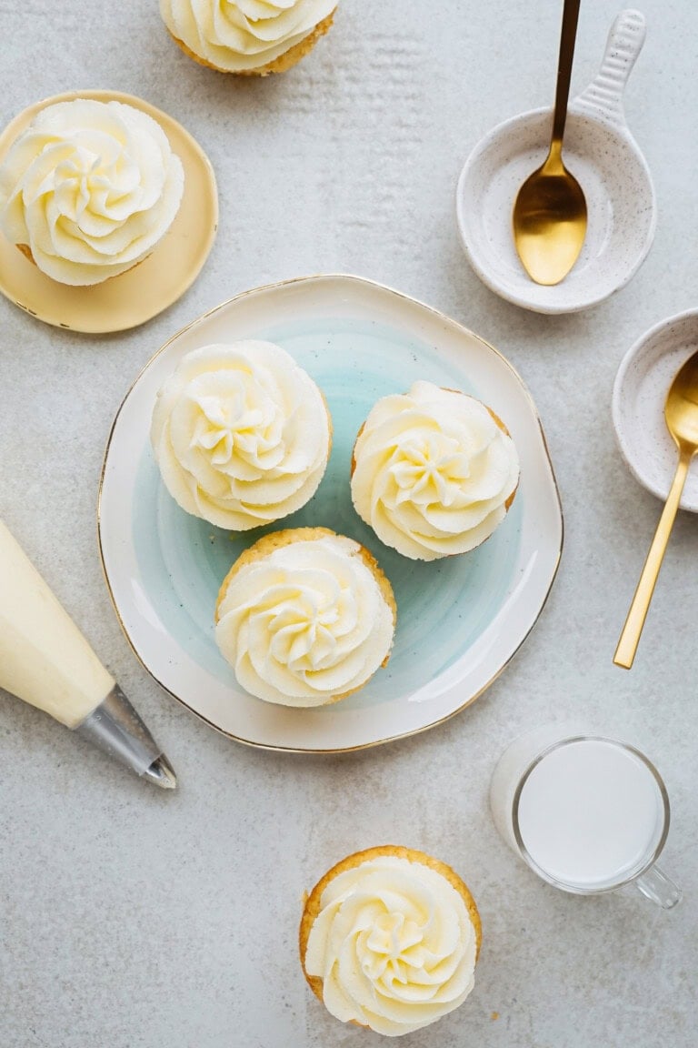 Cupcakes with white frosting on plates, surrounded by gold spoons, small bowls, and a piping bag on a light surface.