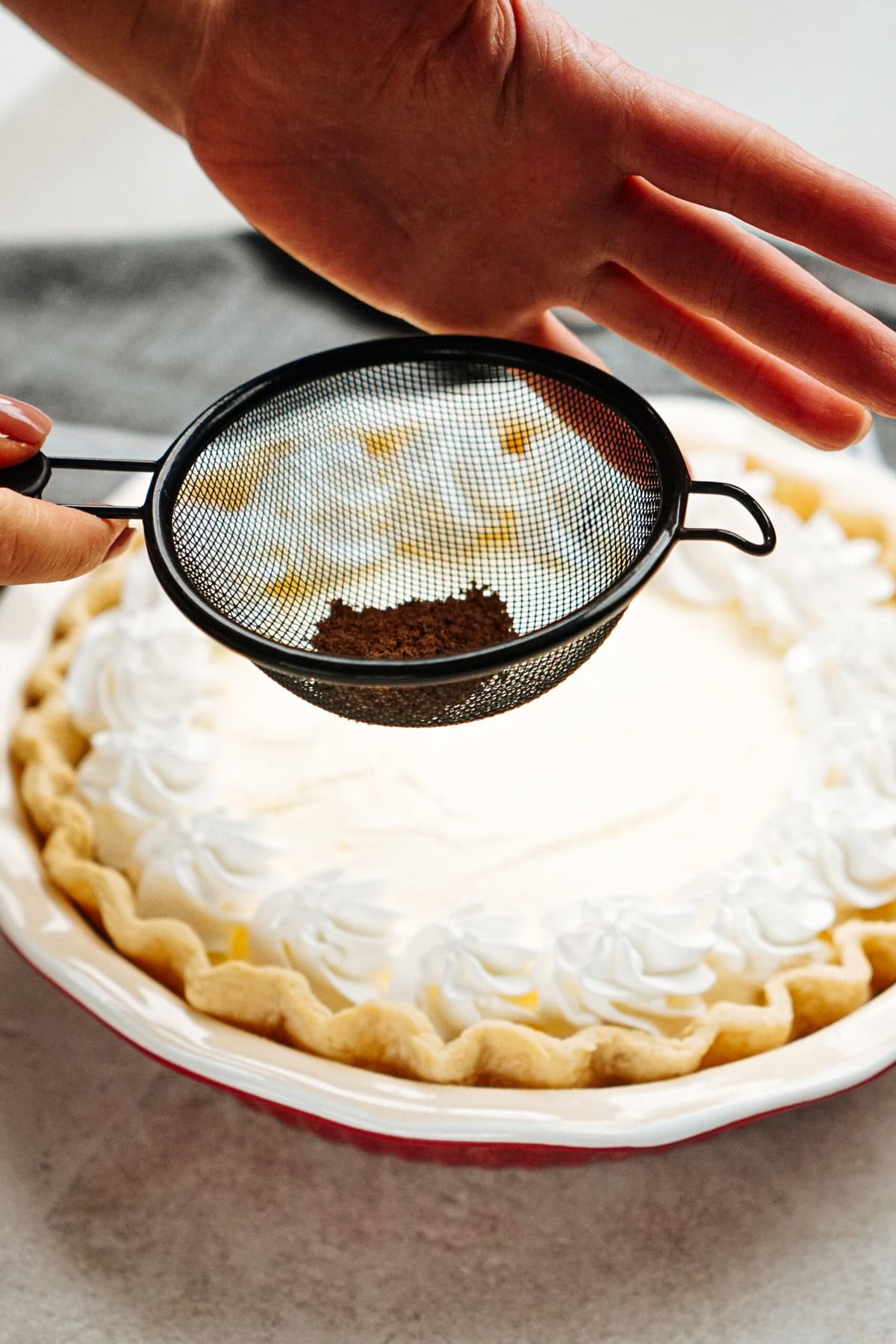 A hand holds a metal sieve dusting cocoa powder over a pie with decorative whipped cream edges.