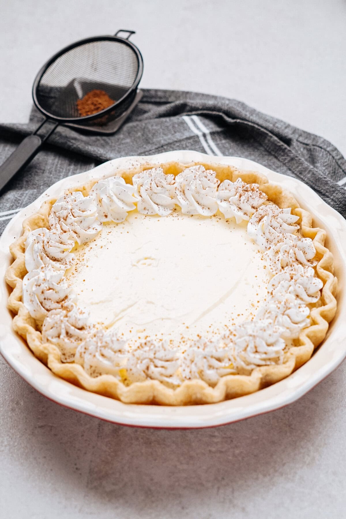 Cream pie with whipped topping and sprinkled cinnamon, set on a gray cloth, with a sieve nearby.