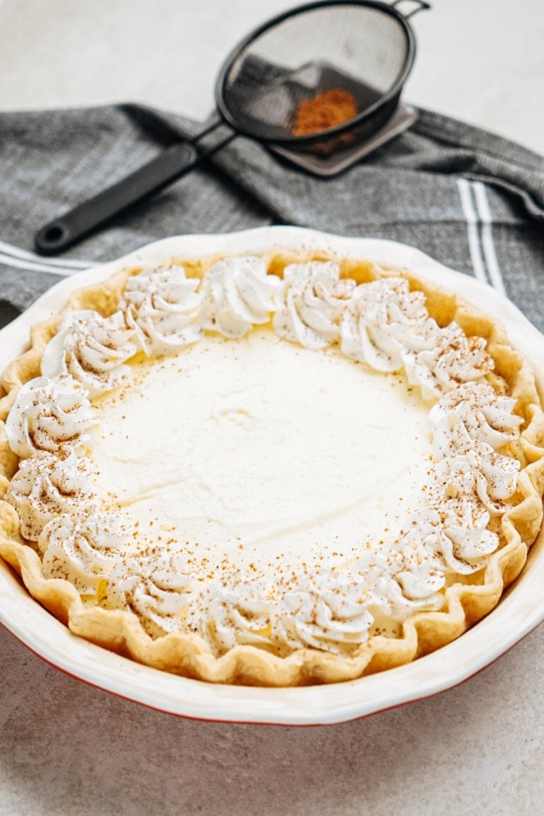 A pie with whipped cream and sprinkled spices on a crimped crust, placed on a white surface with a gray napkin and a sifter in the background.